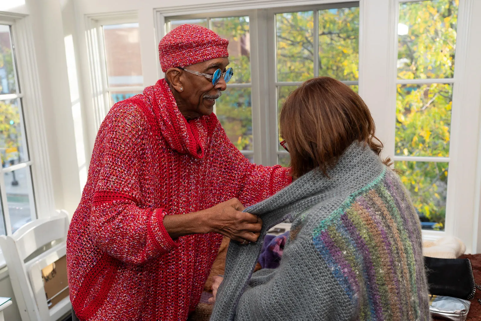 A maker in a red hand-knit sweater wrapping a shawl around a customer by autumn windows