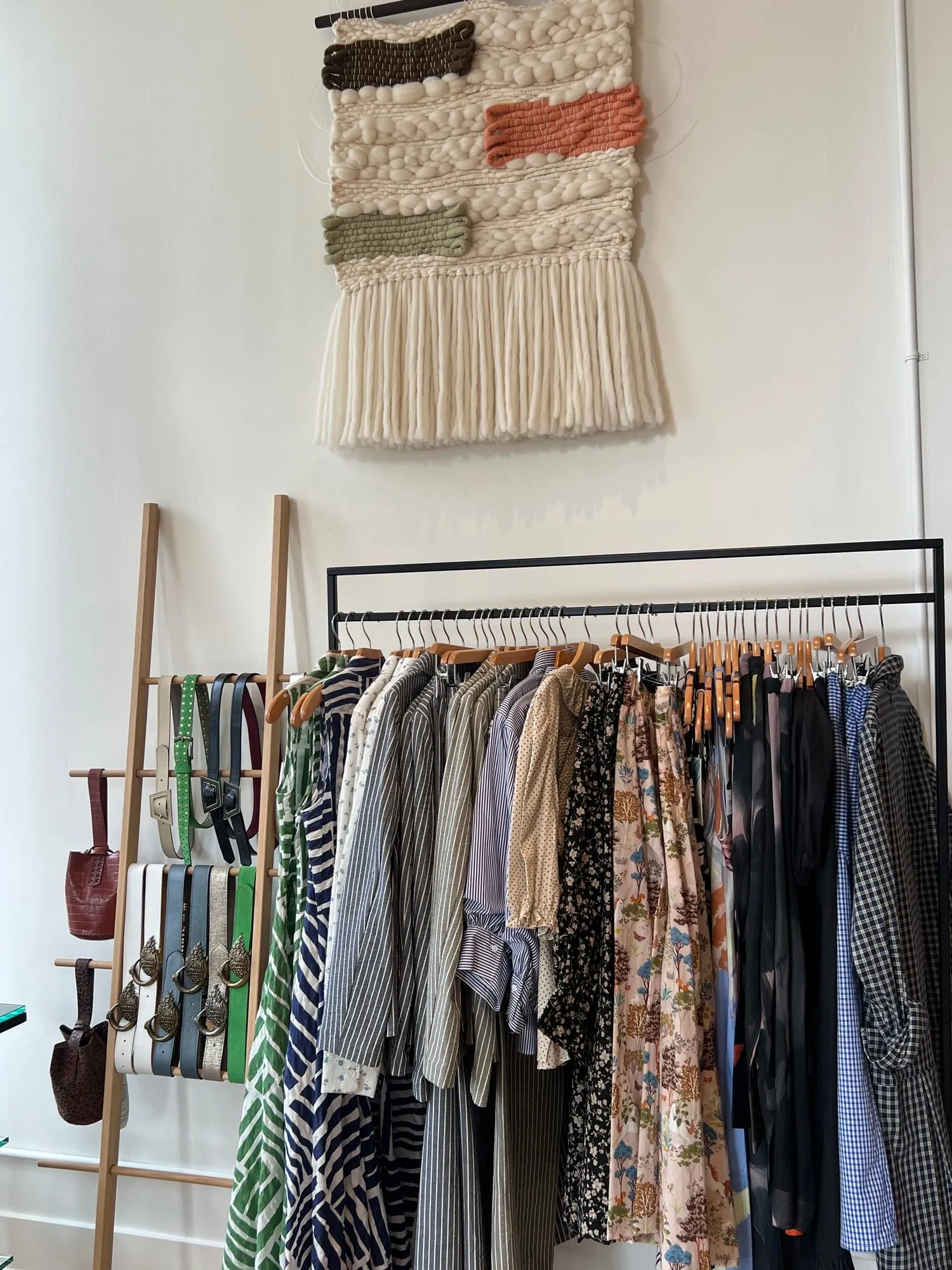 A rack of printed shirts and dresses hanging beneath a hand-woven wall hanging