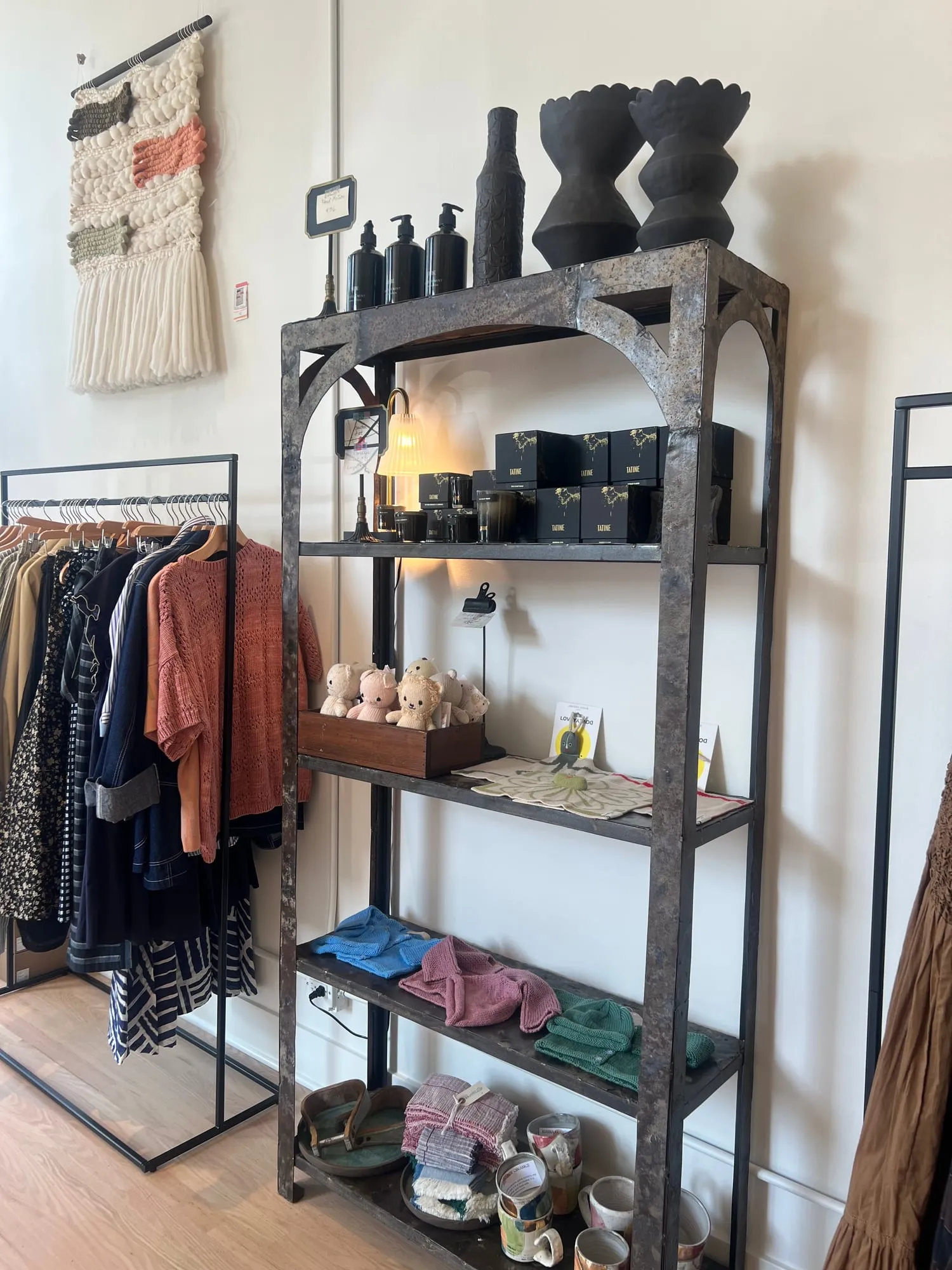 Shelves of candles, boxed goods and stacked ceramics standing between two clothing racks