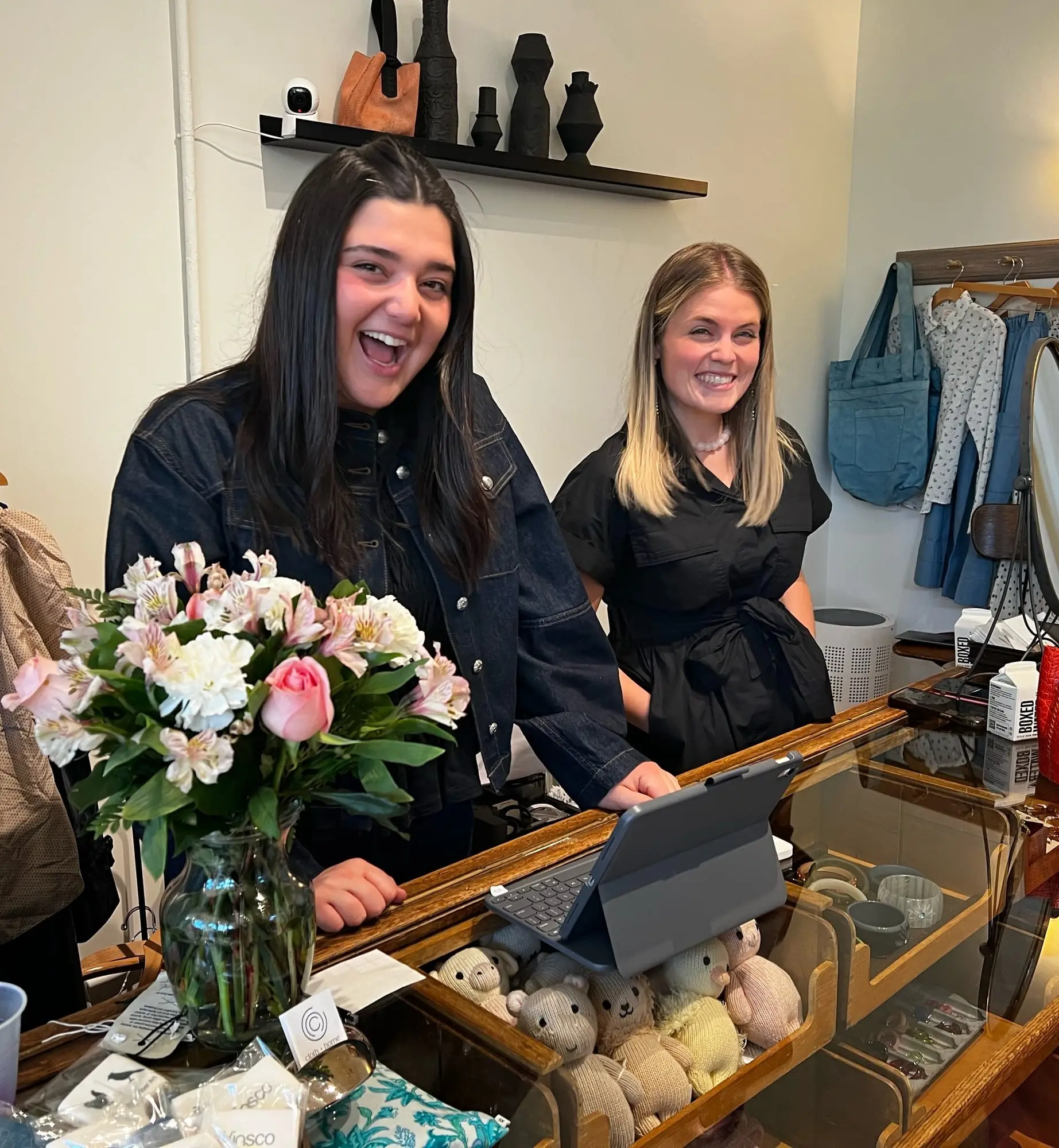 Two members of the cloth + home team laughing behind the shop counter, a vase of pink and white flowers beside them