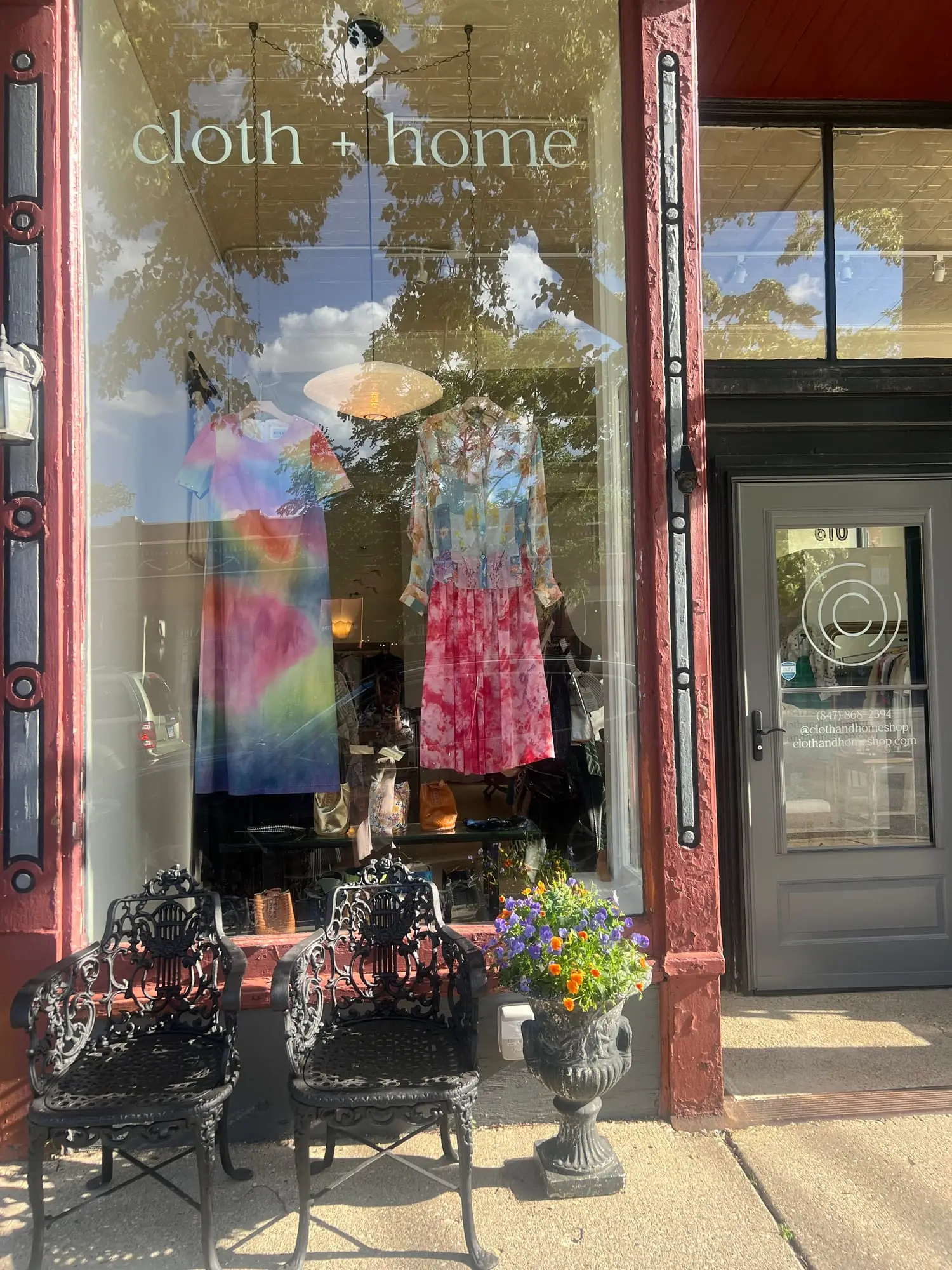 The shop window in afternoon light — hand-dyed dresses hanging behind the glass, a pair of ornate iron chairs and a planter of pansies on the sidewalk