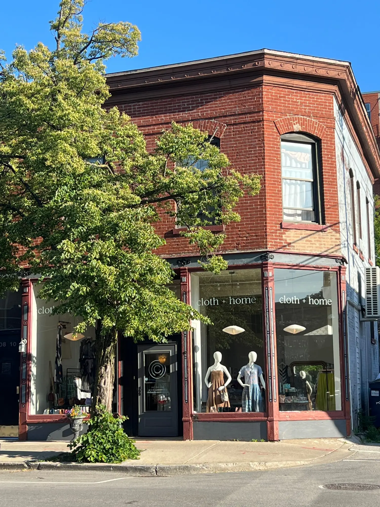 The corner of the cloth + home building on Dempster Street in daylight, red brick under a full summer tree, the name lettered on both windows