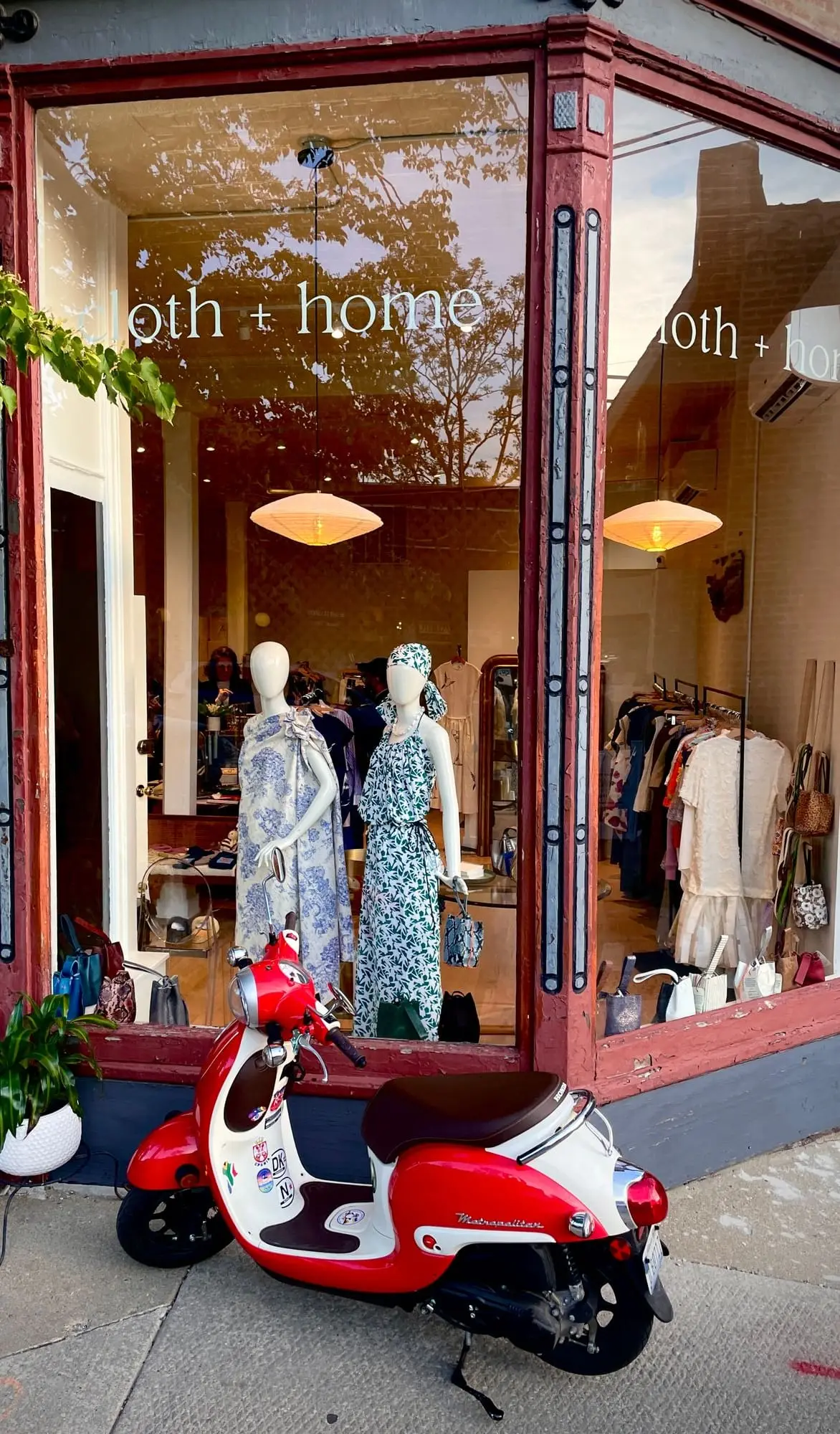 The cloth + home storefront in evening light, a red scooter parked at the kerb and two dressed mannequins in the window behind it