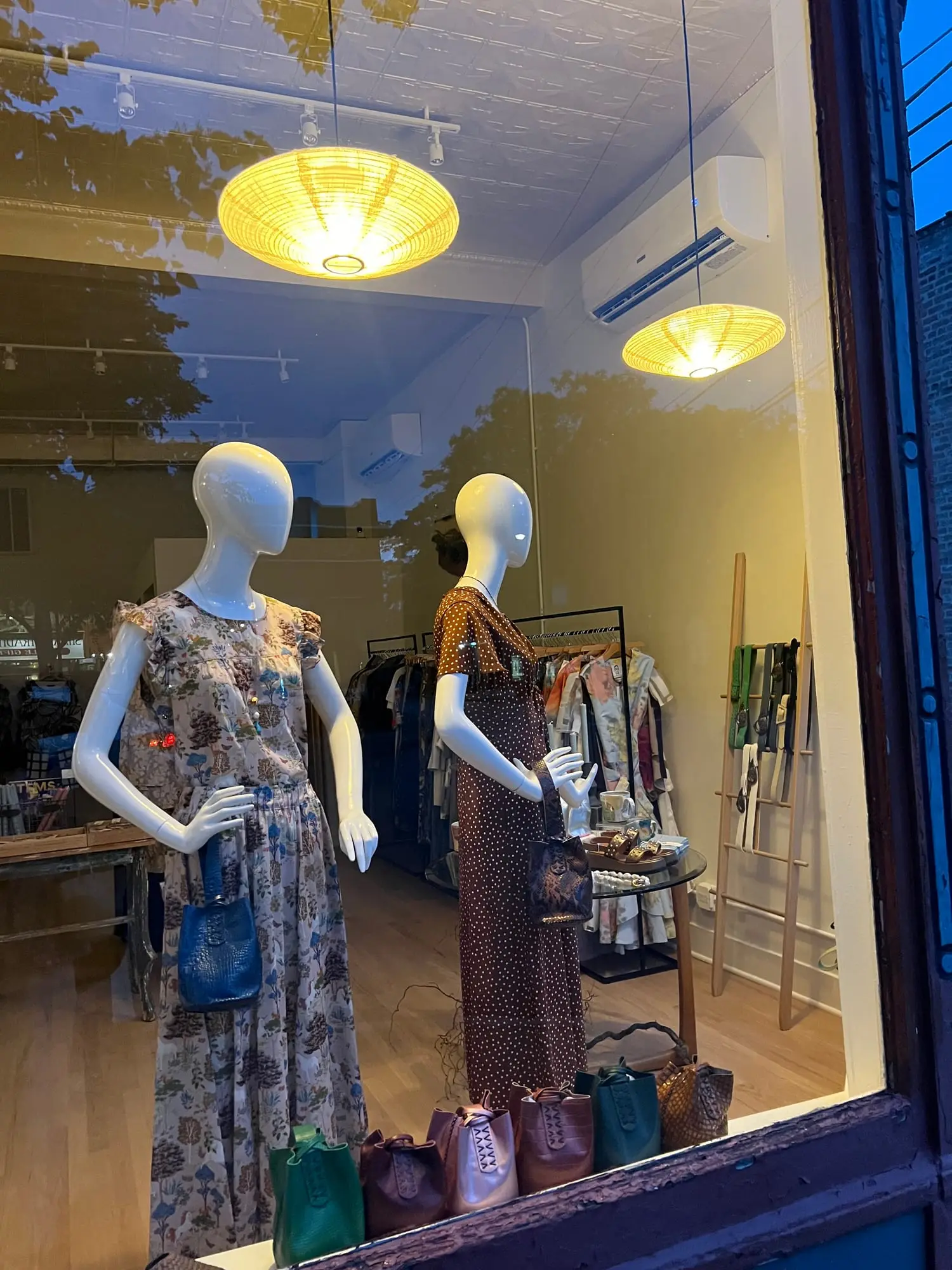 The cloth + home window at dusk — two lit woven pendant lamps over a pair of dressed mannequins, the racks and a row of leather bags visible through the glass