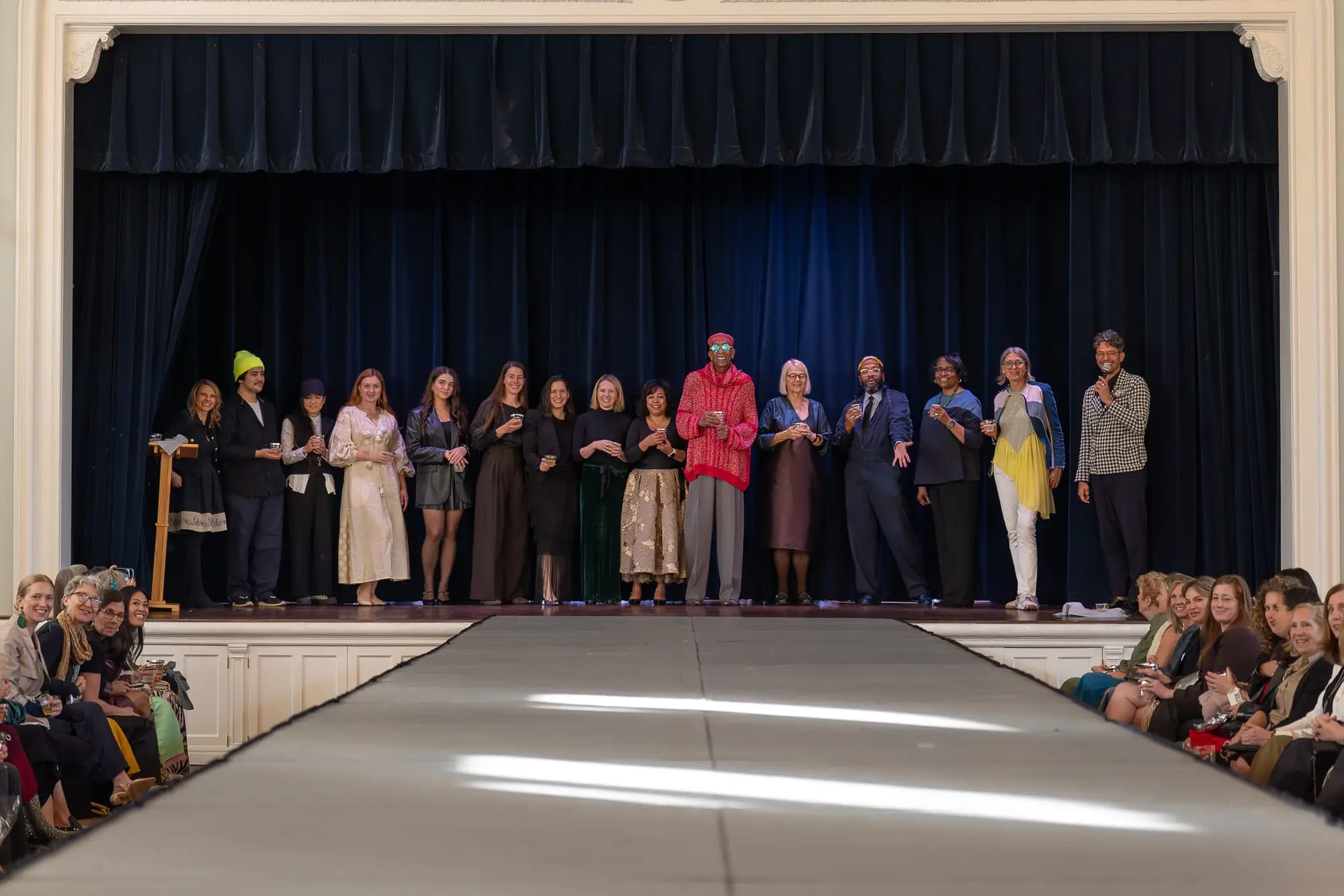The Threaded Stories designers lined up across the stage at the end of the show, raising a toast as the audience looks on