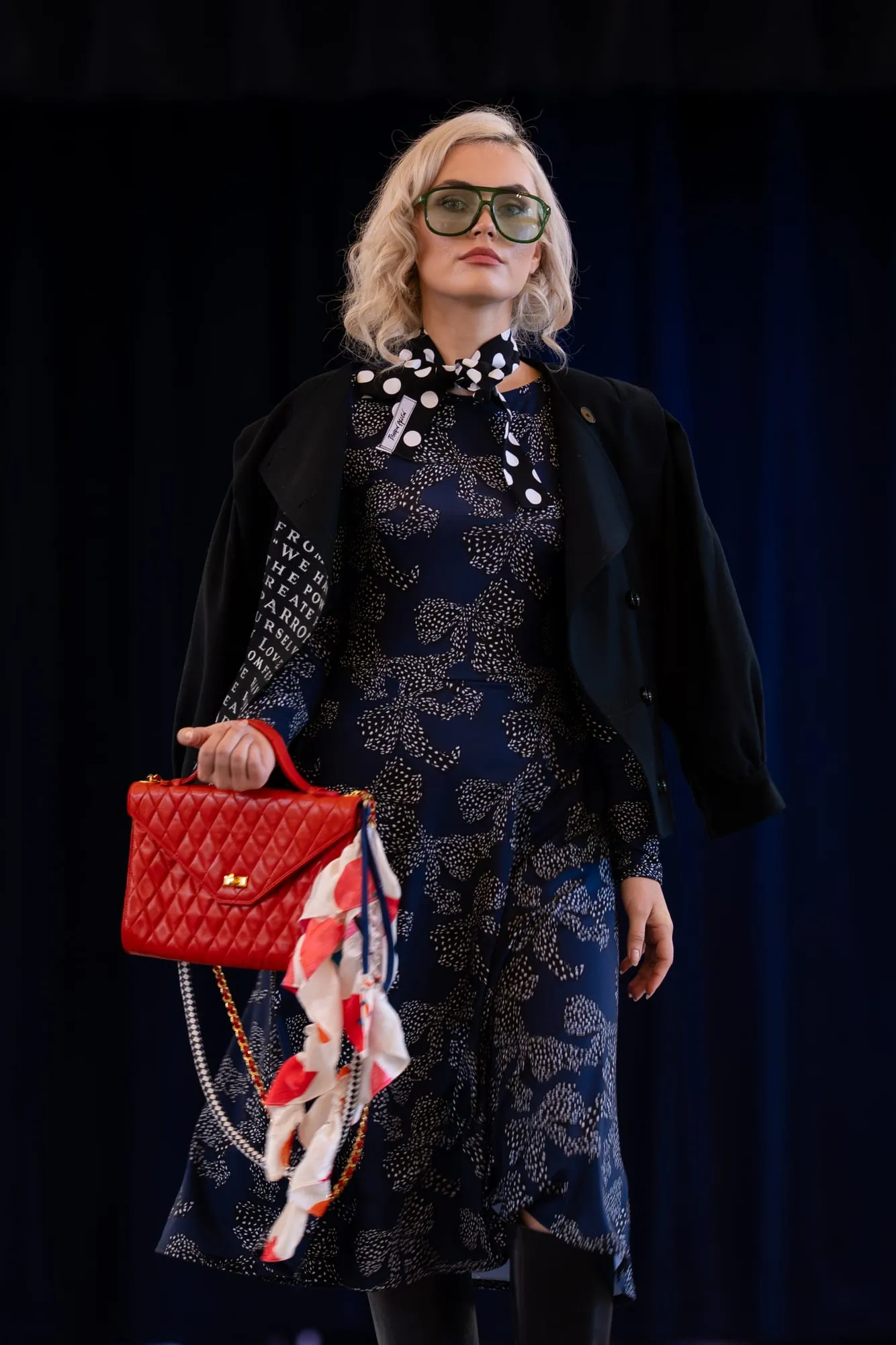 A model on the runway in a navy print dress and black jacket, with a polka-dot scarf and a red bag